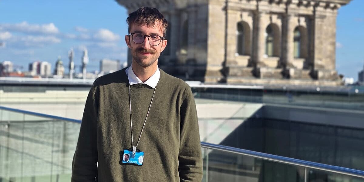 Das Bild zeigt Michael Scharpf vor dem Bundestag in Berlin. Er trägt einen dunkelgrünen Pullover mit darunter liegendem Hemd und hat einen Ausweis umhängen. 