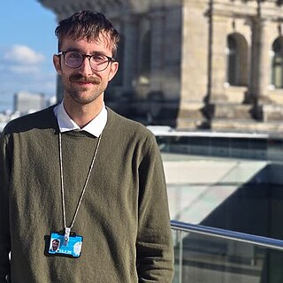 Das Bild zeigt Michael Scharpf vor dem Bundestag in Berlin. Er trägt einen dunkelgrünen Pullover mit darunter liegendem Hemd und hat einen Ausweis umhängen. 