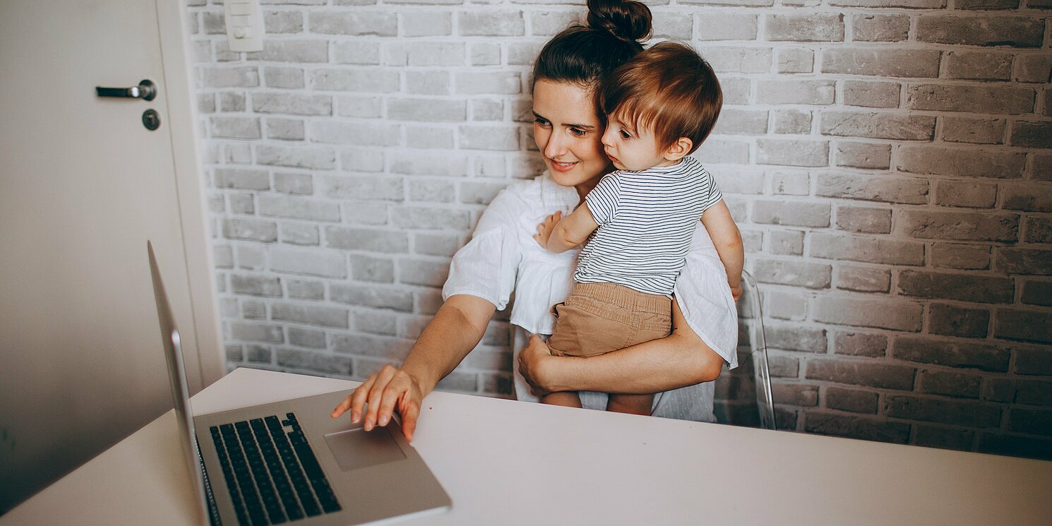 Frau mit Kleinkind auf dem Arm bedient mit der anderen Hand einen Laptop