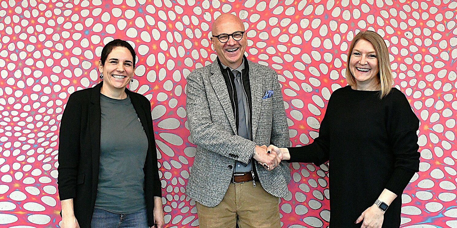 Drei Personen stehen vor einer bunten Wand, ein Mann und eine Frau schütteln sich die Hand.